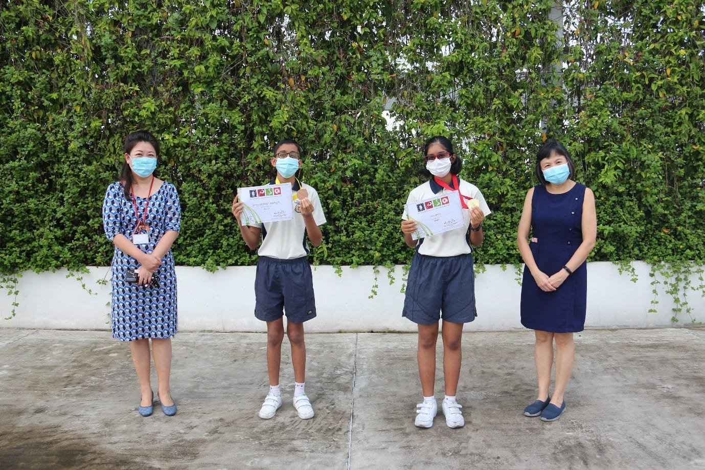 Four people wearing face masks stand outdoors; two students hold certificates and medals beside two adults.